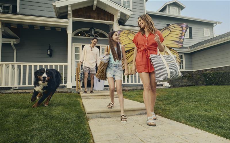 Woman in red dress with yellow butterfly wings and young girl walk out of house smiling at each other with large bags, a dog, and a man following behind.