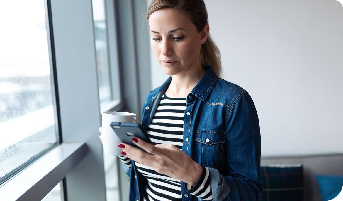 Woman in denim jacket and striped shirt stands by a window with a mug in one hand and looking down at her phone in the other hand
